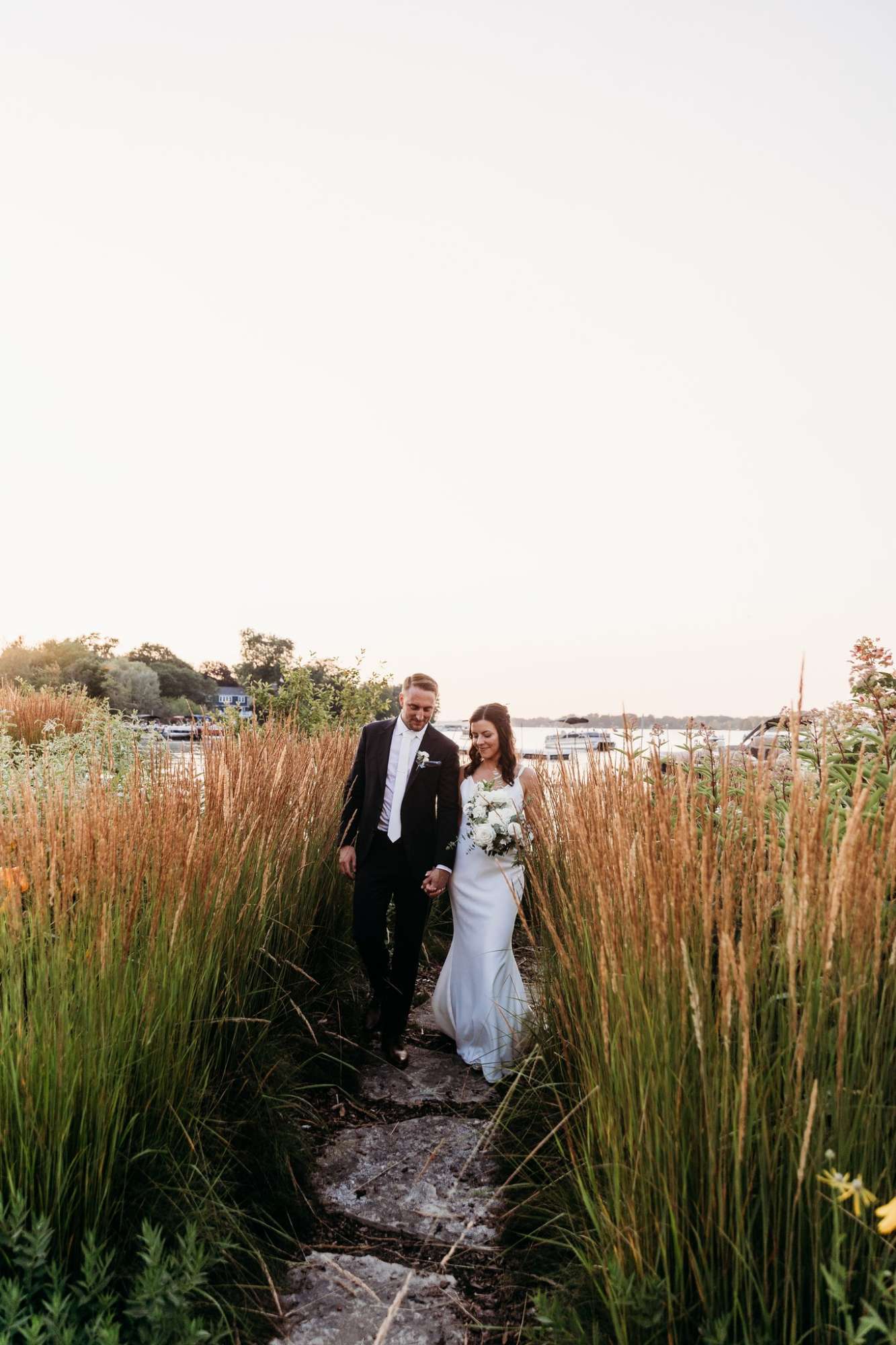 Bride and groom walking hand in hand through tall grass at sunset with lake in the background at Oconomowoc Community Center.