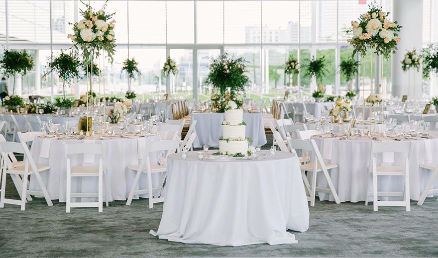 White linens, table candles, high centerpiece floral arrangements with lots of greenery plus cake table in forefront.Wedding reception in white at Bartolotta Events & Catering at Discovery World
