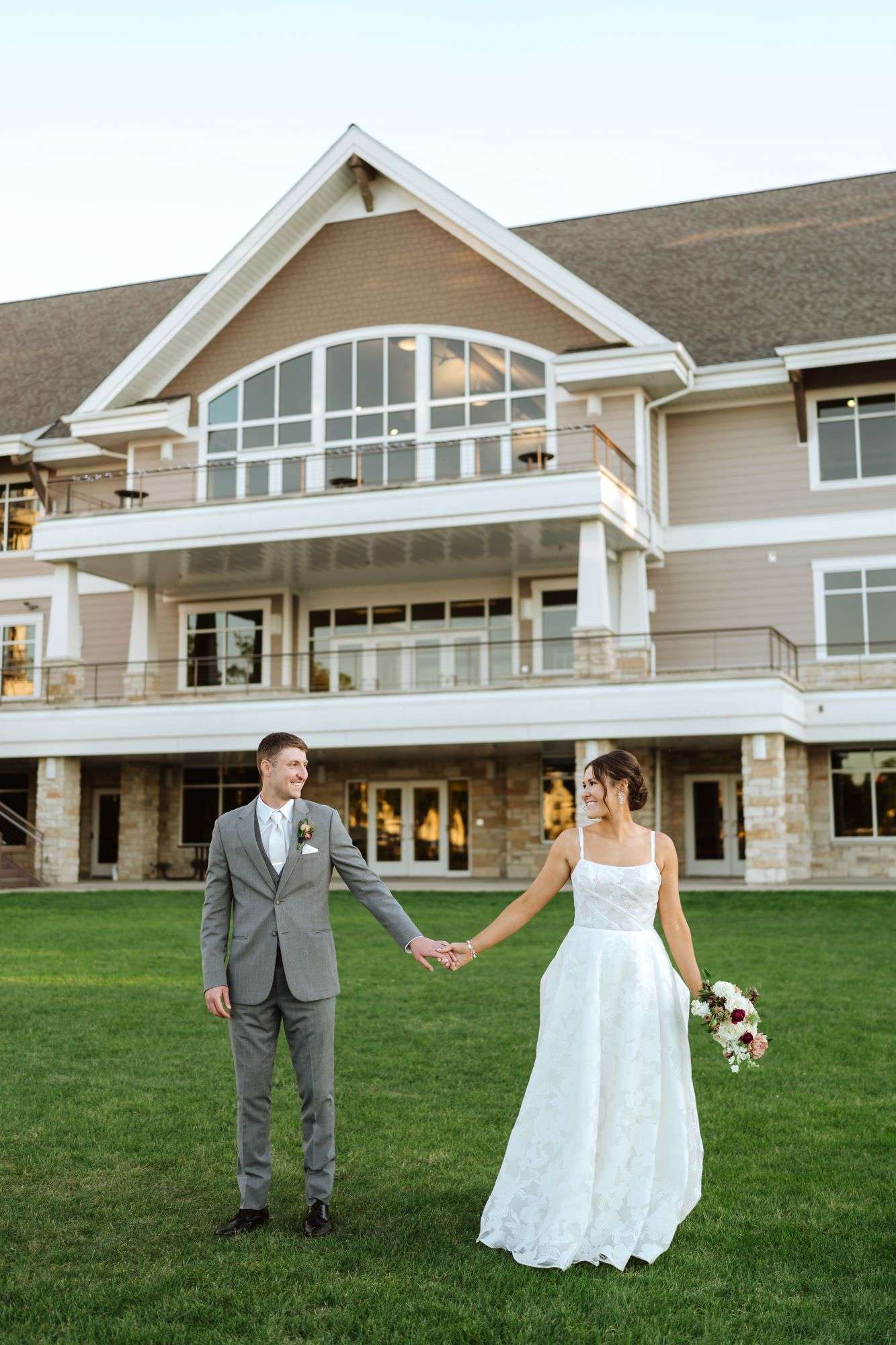 Bride and groom holding hands on a lawn in front of Oconomowoc Community Center, a modern event venue with large windows and wrap-around balcony.