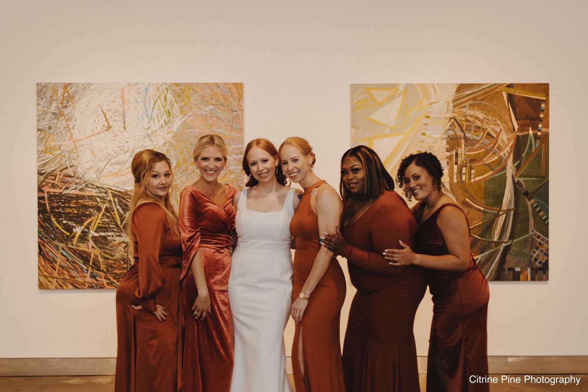 Bridesmaids in burnt orange dresses pose with the bride in Jan Serr Studio's art-filled gallery space.