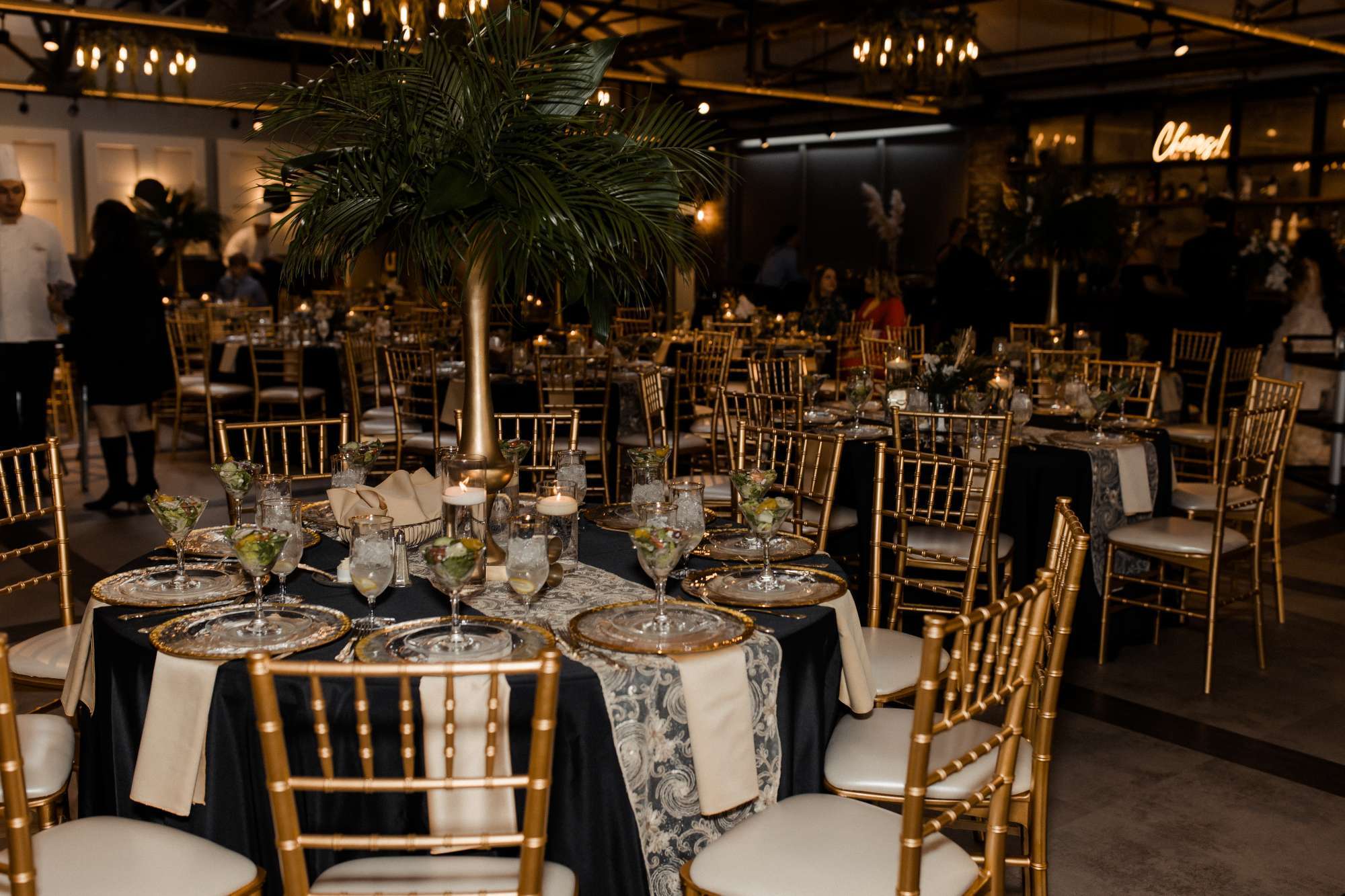 A glamorous reception space styled by Ambrosia Events features golden chairs, black linens, and dramatic centerpieces with lush palm leaves for a modern, upscale look.