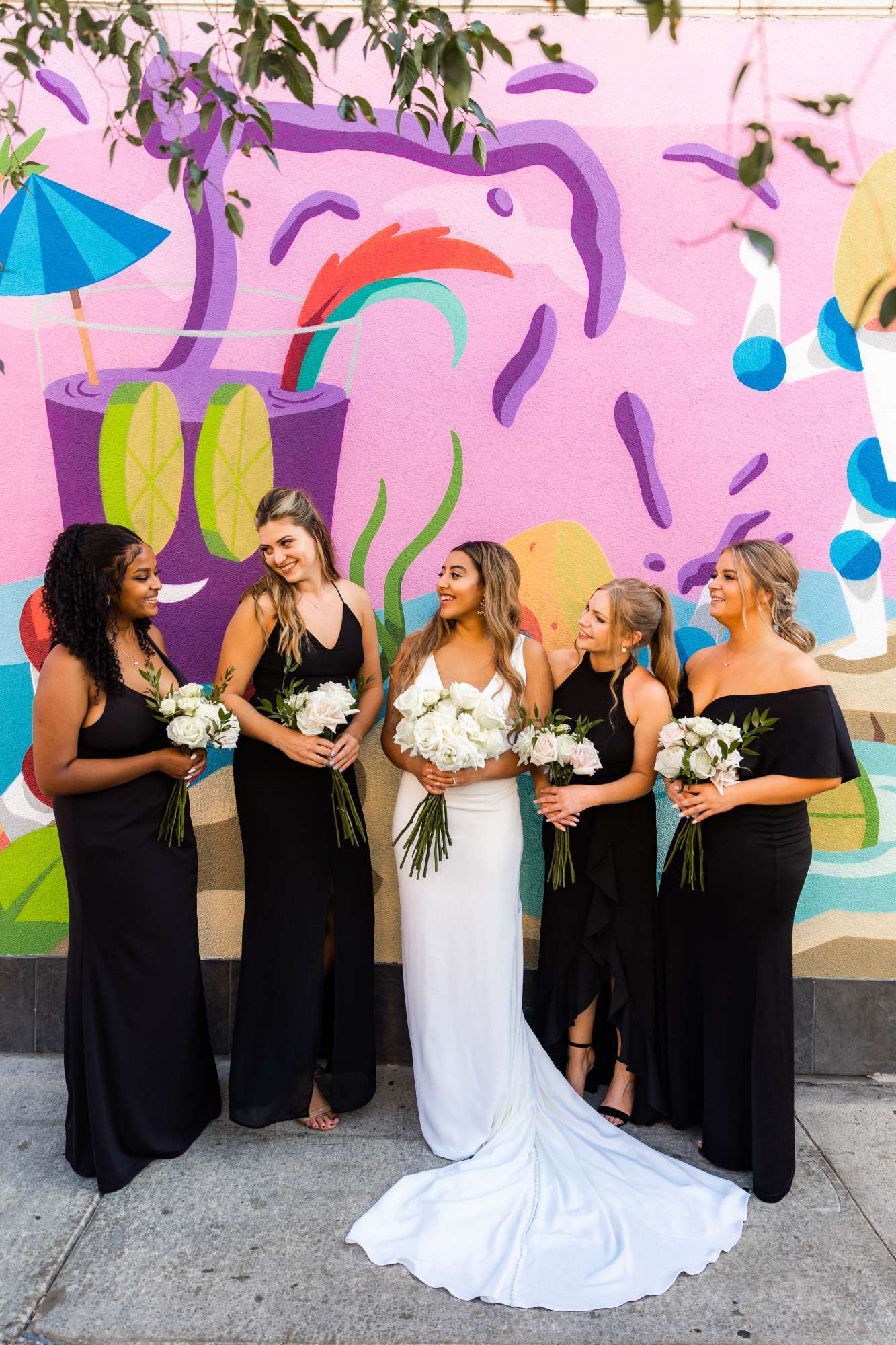 A chic bridal party styled by Ambrosia Events poses against a colorful mural, holding elegant white floral bouquets.