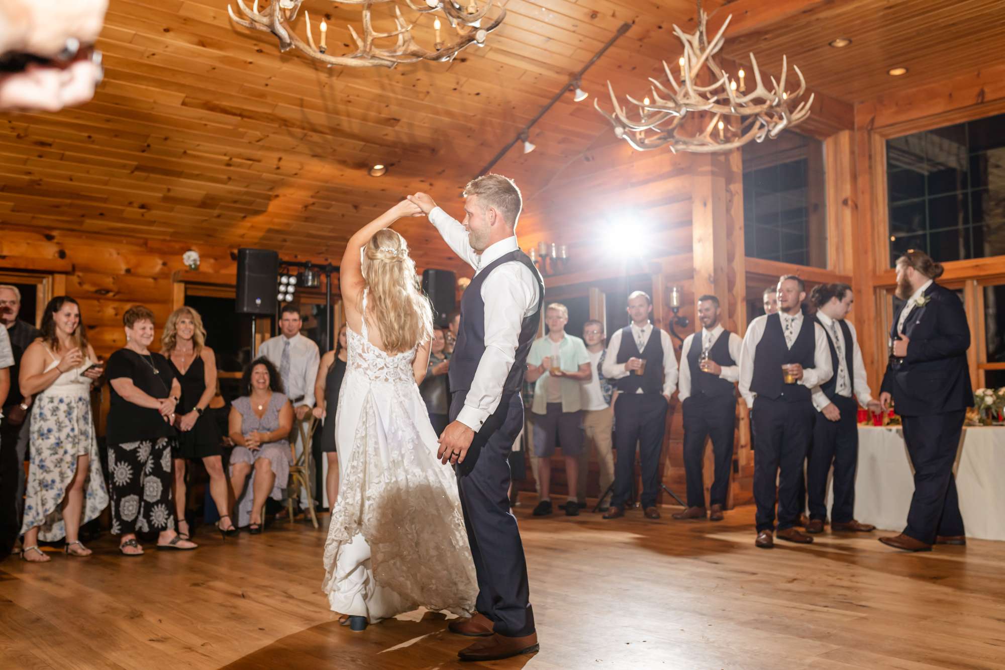 The bride and groom share a romantic first dance inside Milford Hills' rustic reception hall, surrounded by friends and family under warm antler chandeliers.