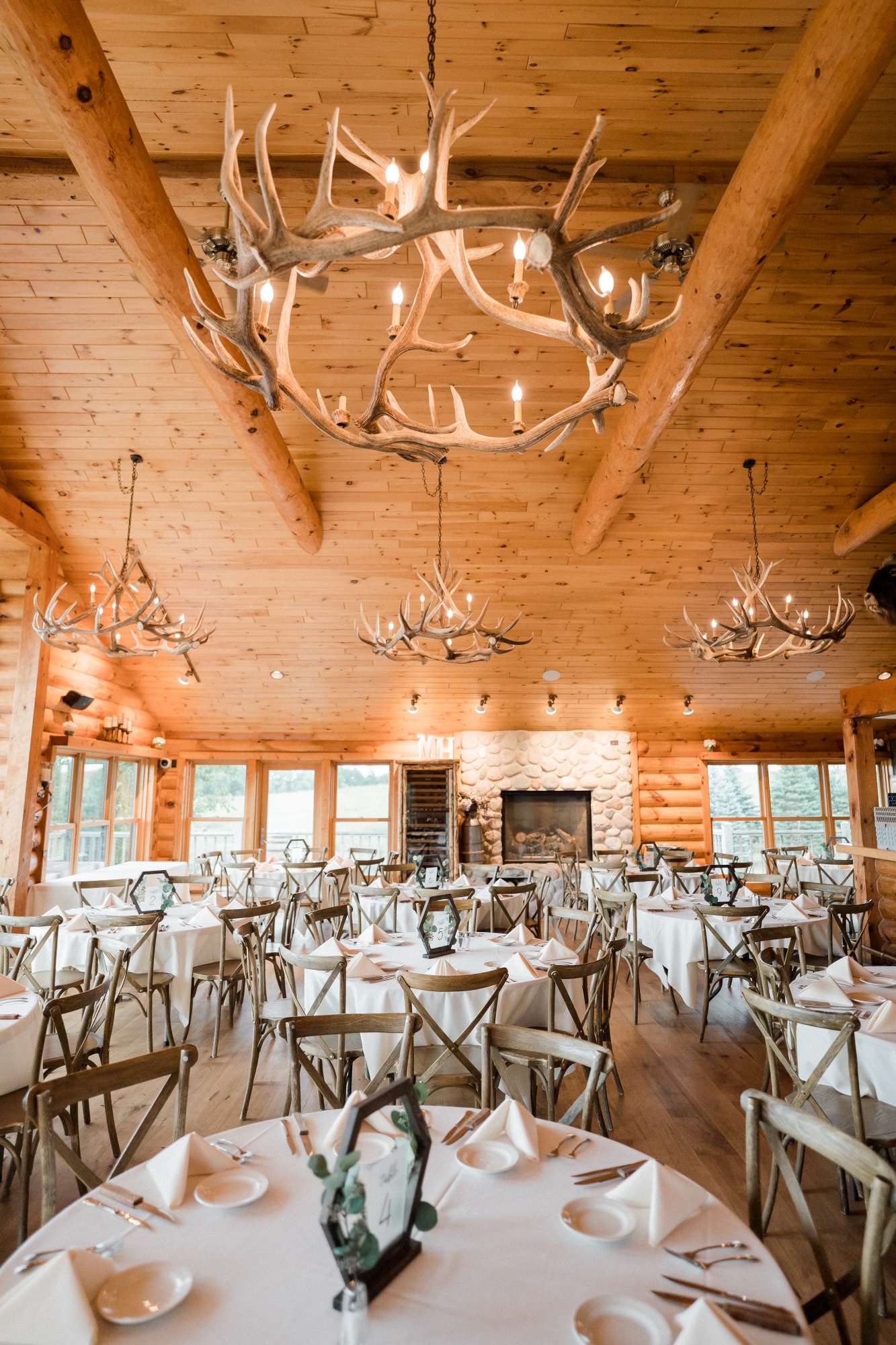 A wide-angle view of the warmly lit Milford Hills reception hall, showcasing rustic wooden beams, stone fireplaces, and elegantly arranged tables.
