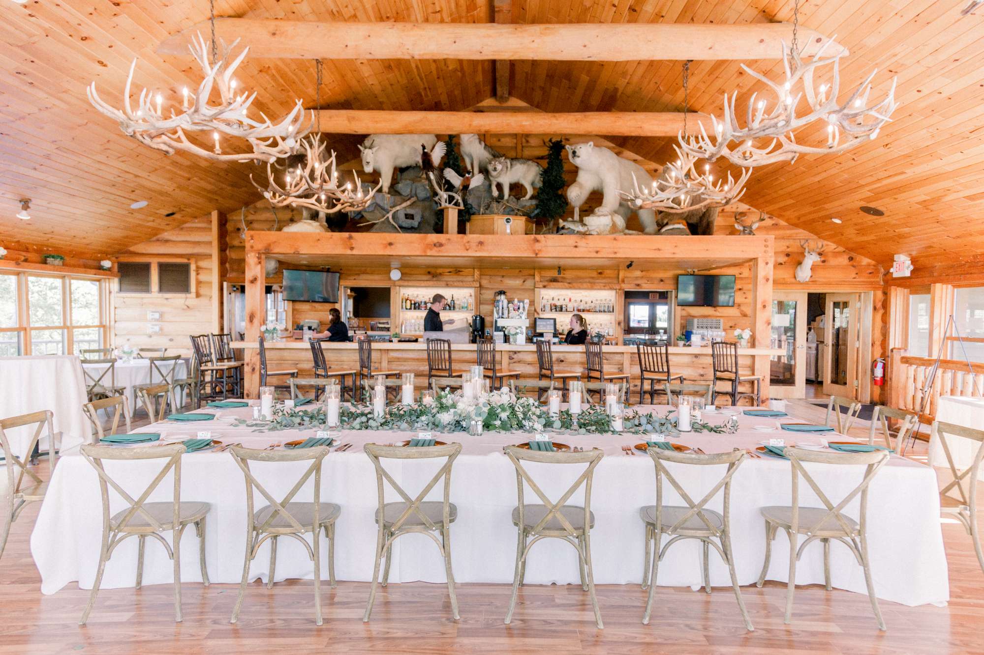 A breathtaking reception setup at Milford Hills, with long banquet tables adorned with lush greenery, candlelight, and antler chandeliers beneath an impressive wooden ceiling.