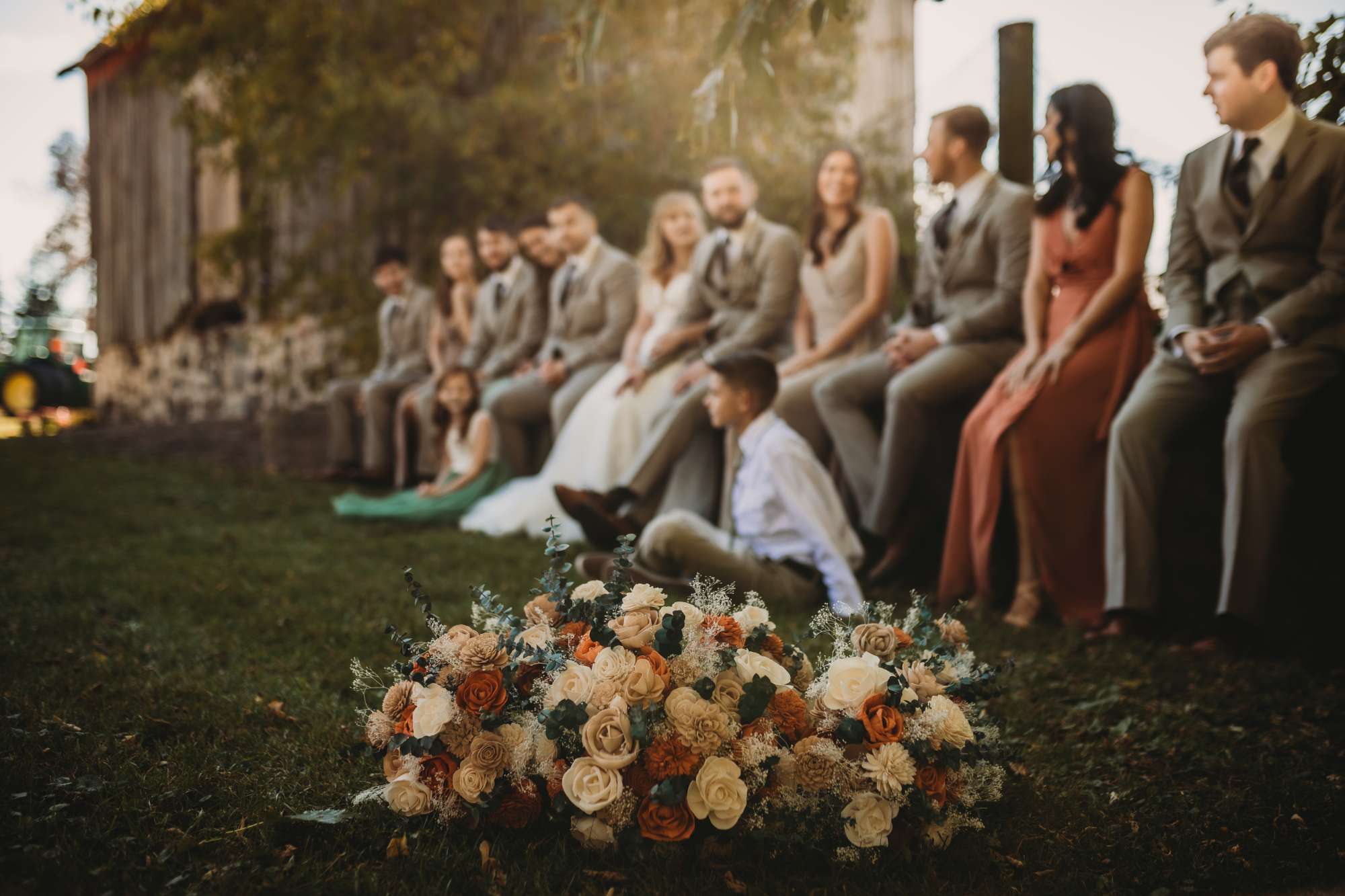 A wedding bouquet with warm, earthy tones rests on the grass in front of a blurred background of the wedding party at Milford Hills.