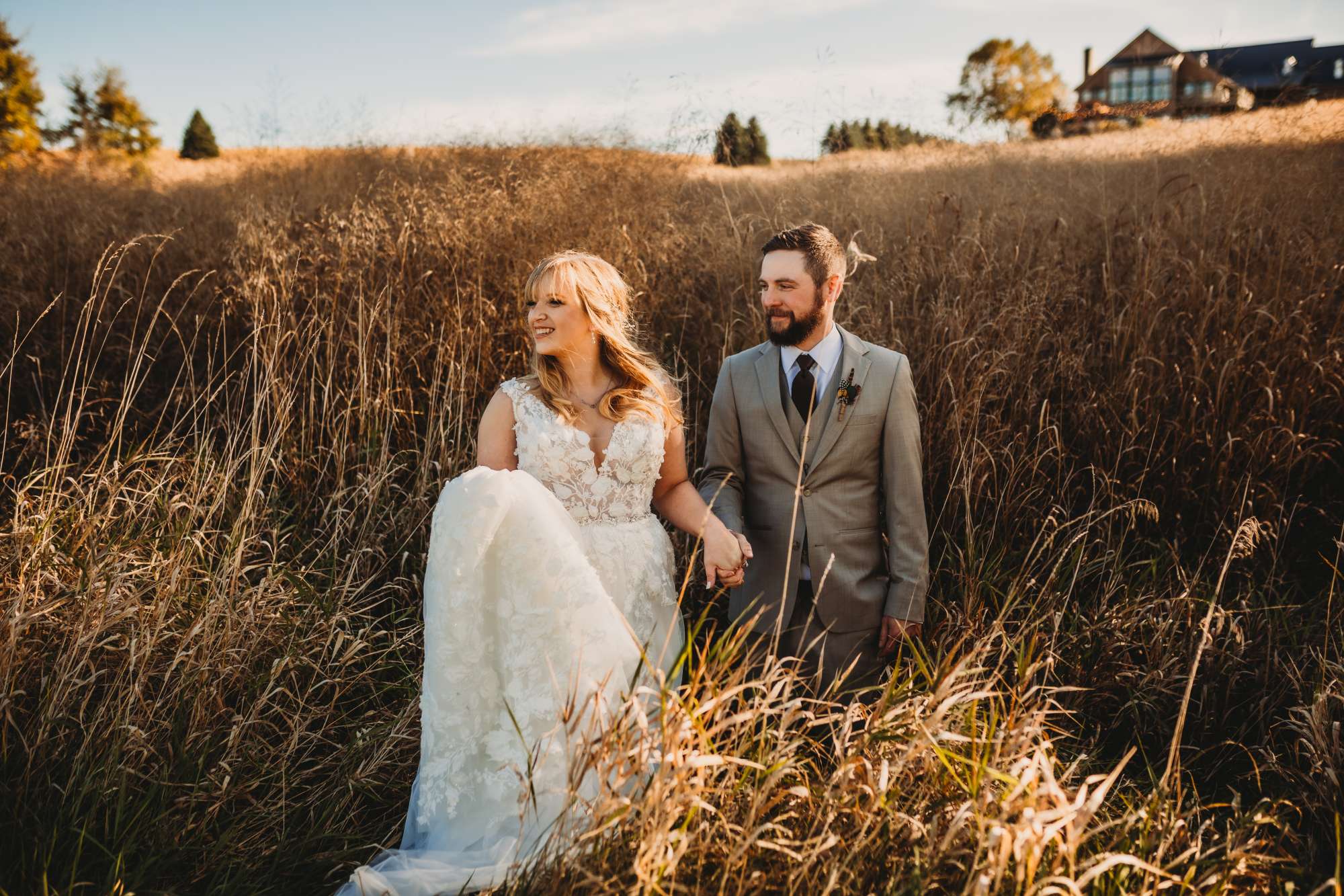 The newlyweds wander through tall grass at Milford Hills, with the rustic lodge in the background, basking in the golden sunlight of a perfect wedding day.