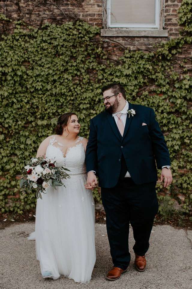 Beautiful bride with bouquet and groom near brick wall covered in ivy.