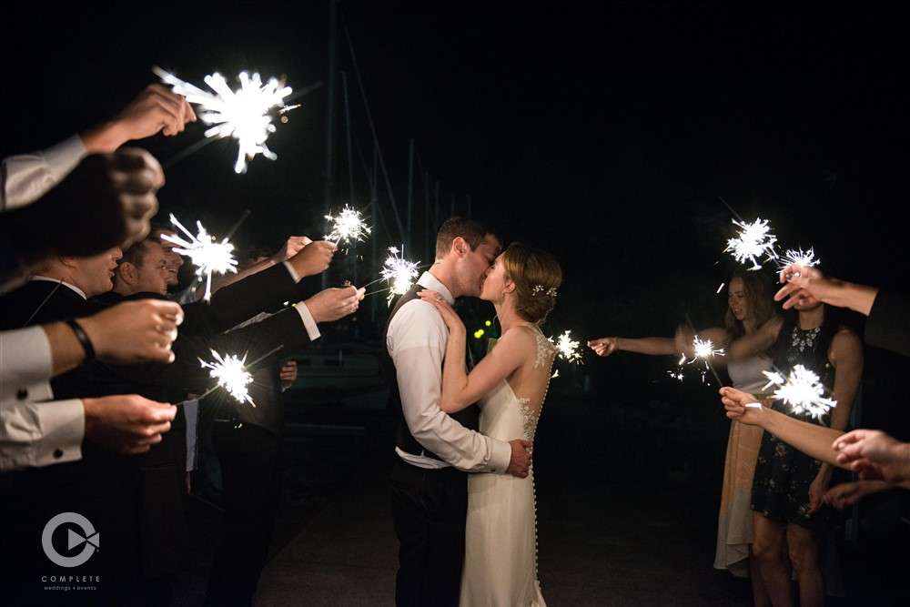 Wedding party waves sparklers as bride & groom kiss