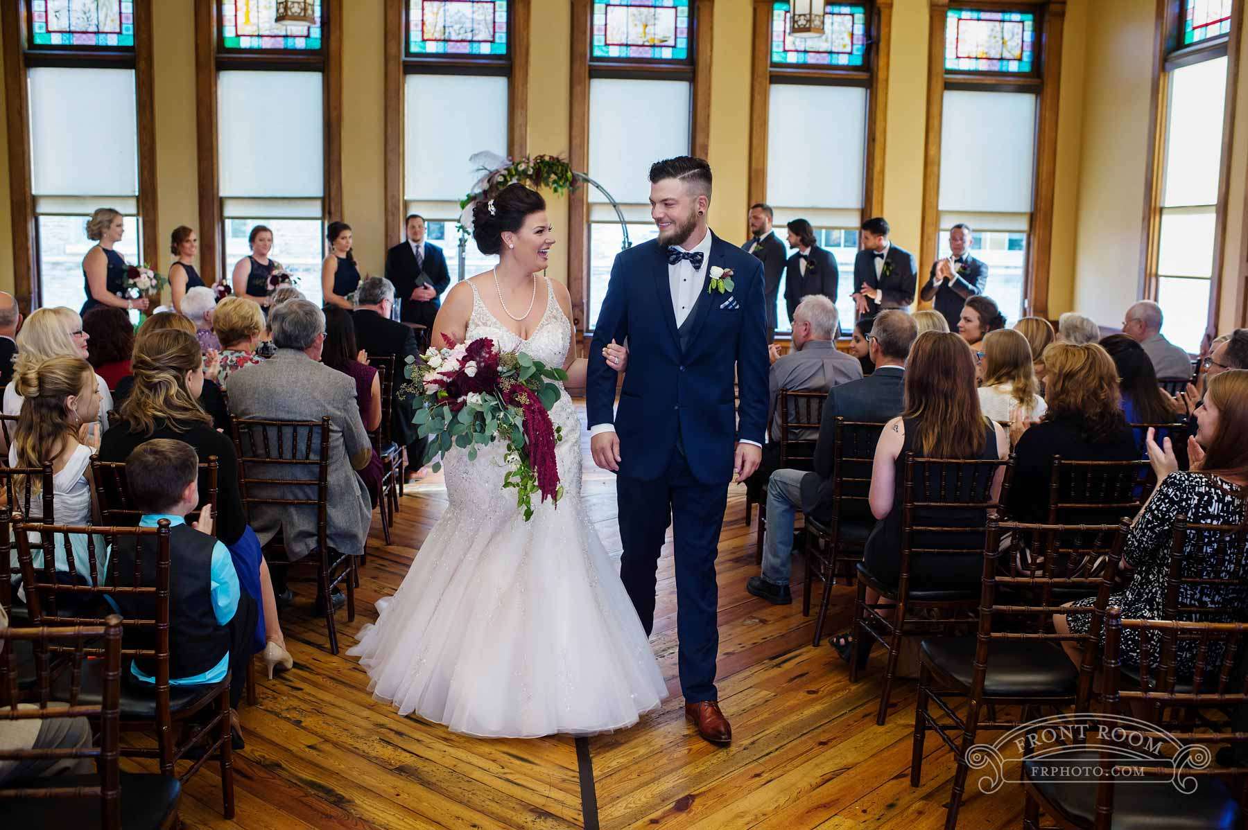 A beaming couple walks down the aisle in the historic Great Hall surrounded by family and friends at Best Place at Historic Pabst Brewery.