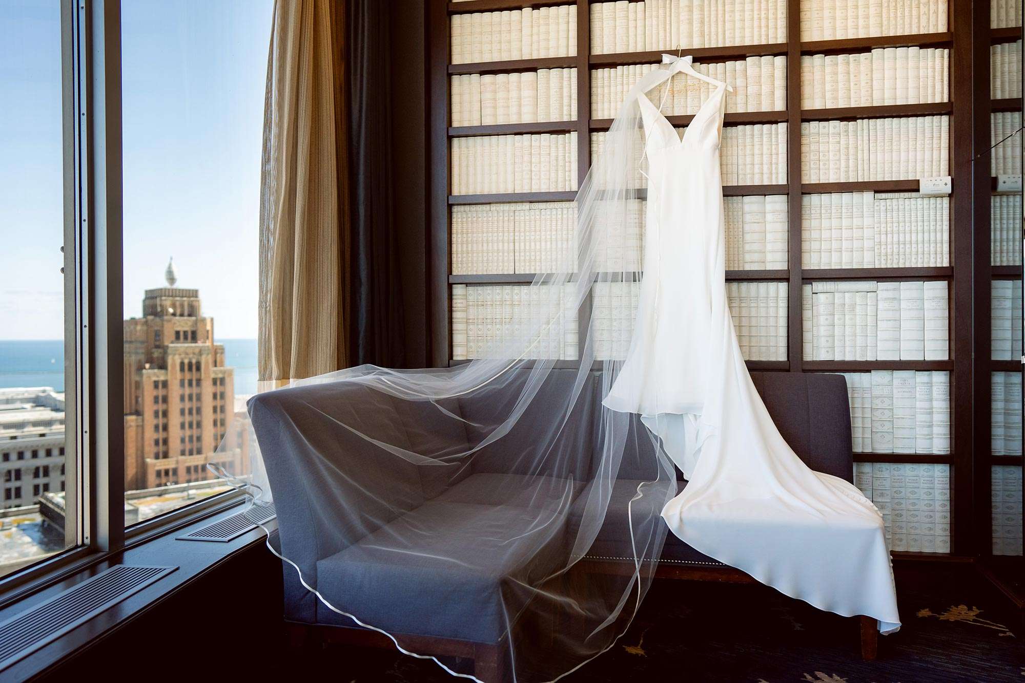 Bridal gown from White Dress hanging against cream books on wooden shelving with veil splayed out on corner sofa at the Pfister