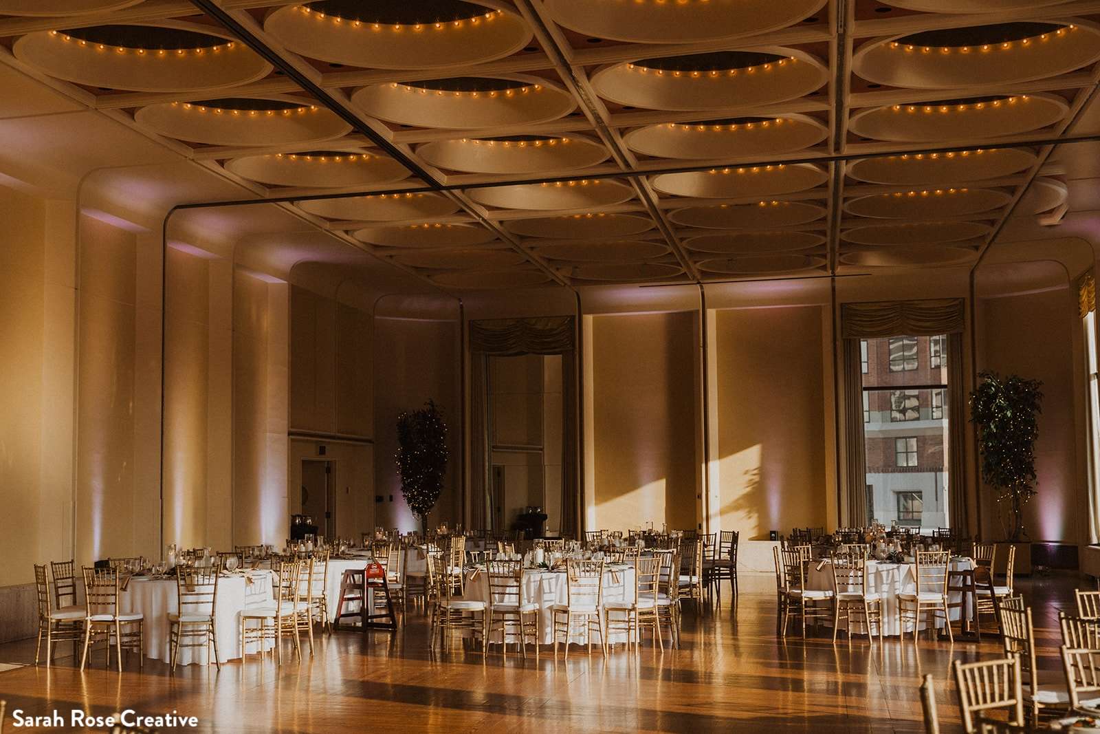 The grand ballroom at Marcus Performing Arts Center, set with round tables, gold chairs, and lit with golden hour sunlight streaming through large windows.