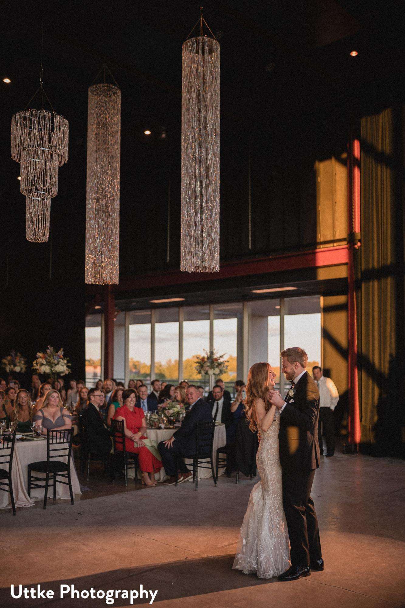 A newlywed couple shares their first dance under elegant chandeliers at Jan Serr Studio, surrounded by seated guests in a warmly lit reception hall.