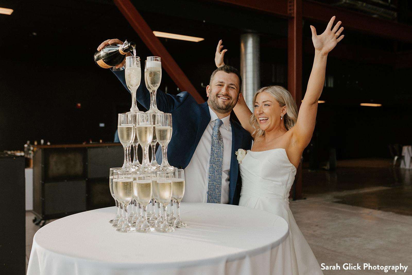 A bride and groom celebrate with a champagne tower at Jan Serr Studio, pouring bubbly and cheering with joy in a modern event space.