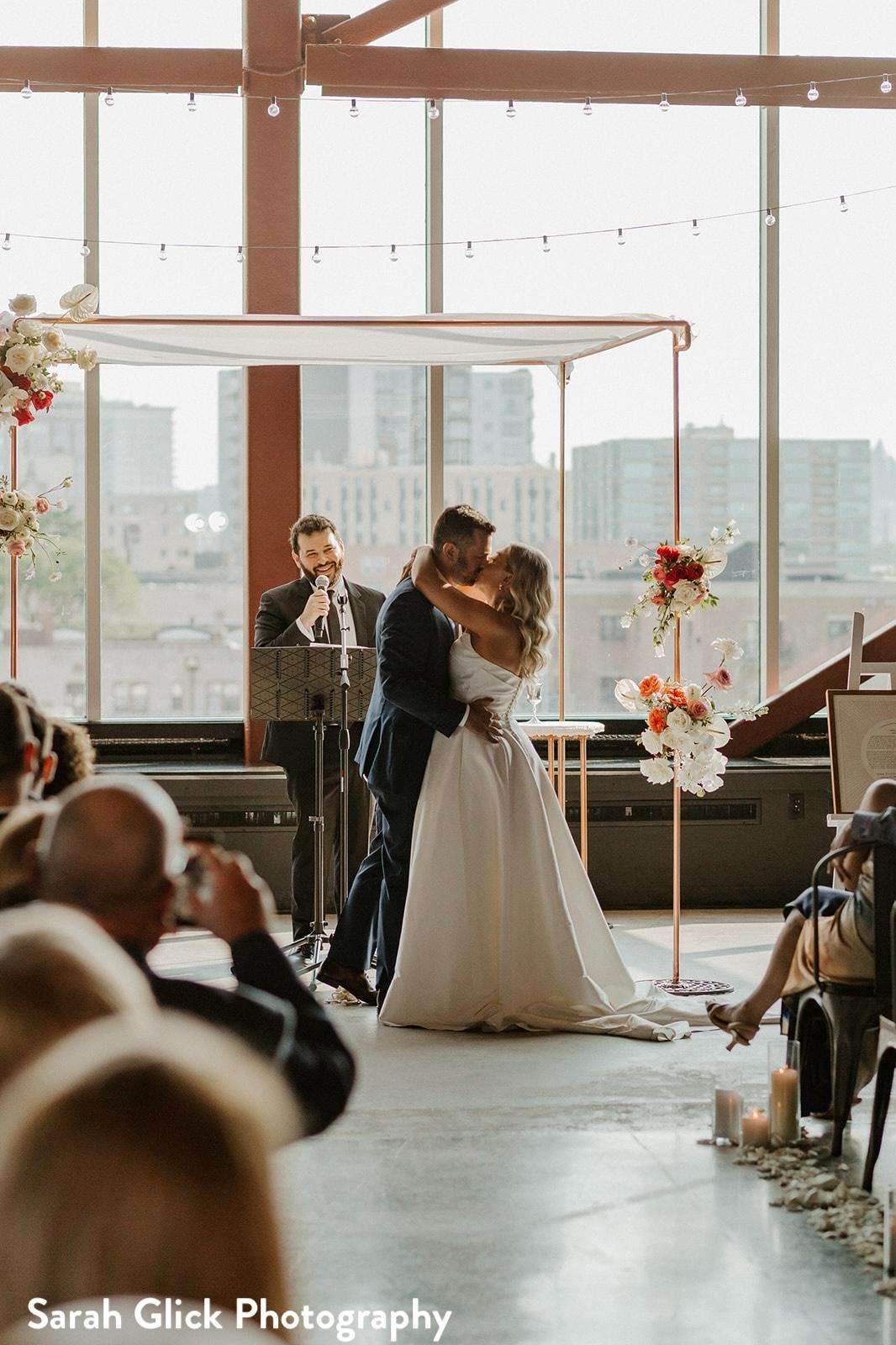 A couple exchanges vows under a minimalistic arch adorned with flowers at Jan Serr Studio, set against panoramic city views.