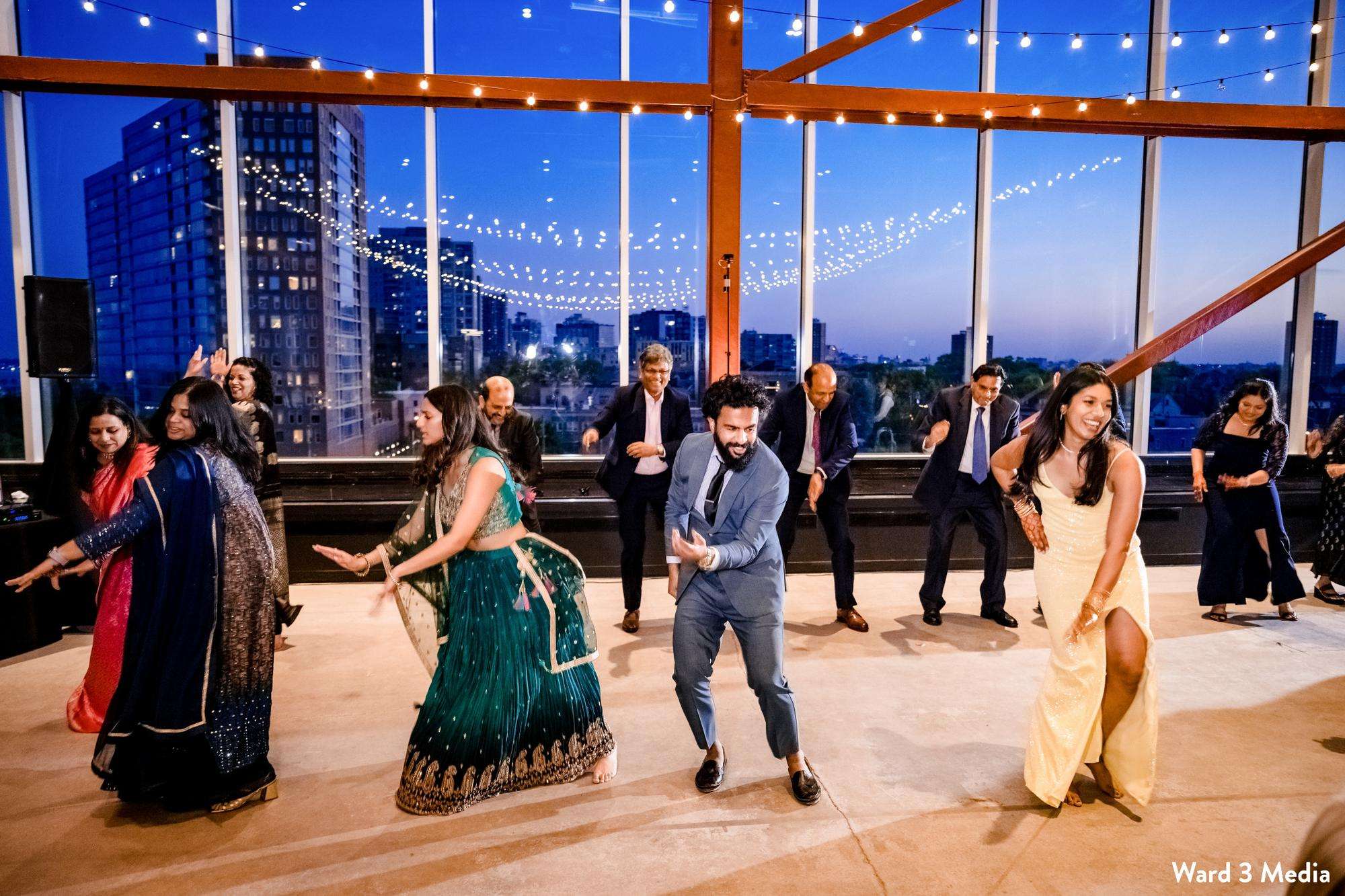 Guests engage in energetic dancing during a festive evening reception at Jan Serr Studio, framed by string lights and large windows.