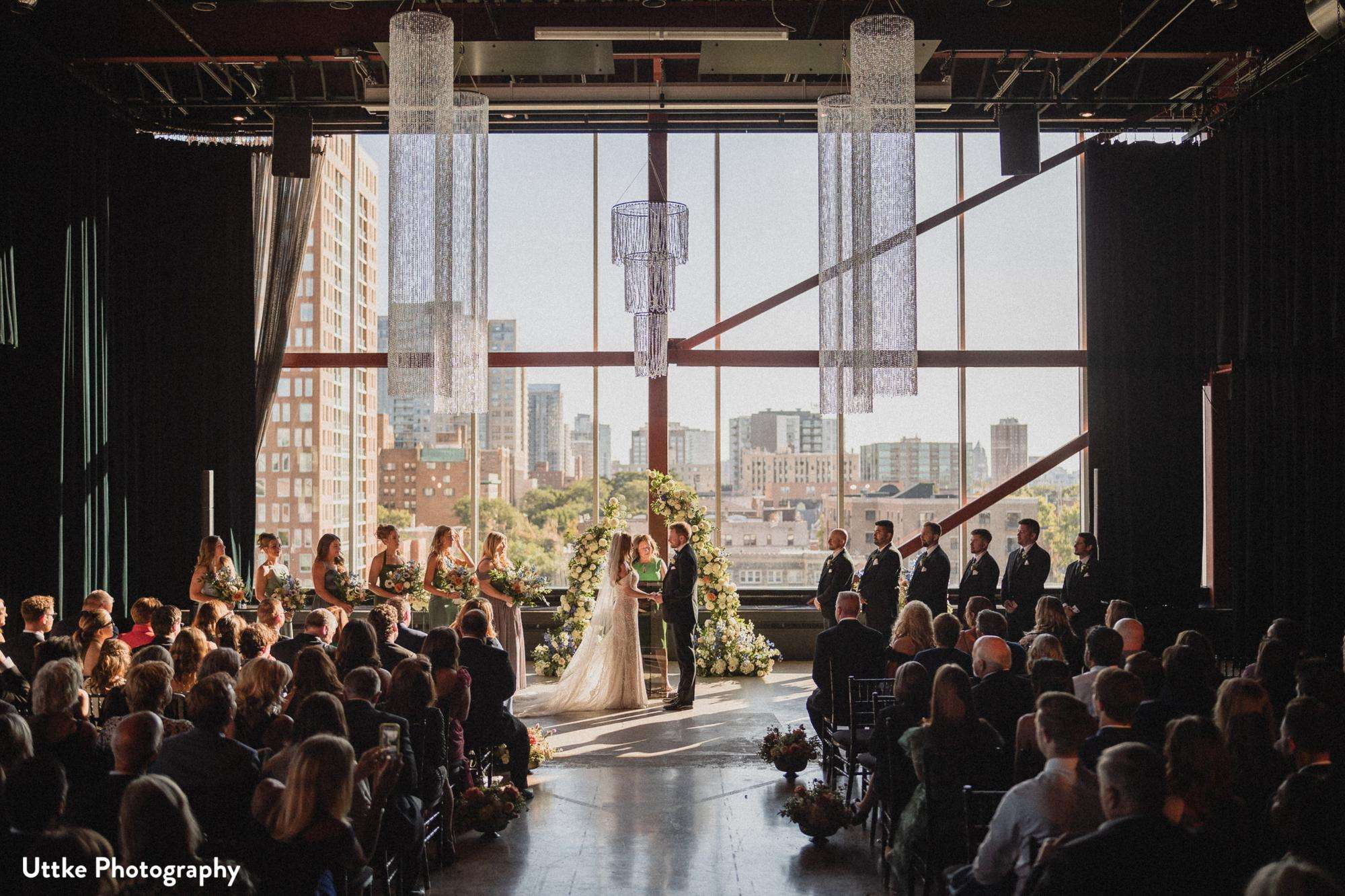 A wedding ceremony scene at Jan Serr Studio with a couple exchanging vows in front of a large audience, framed by elegant chandeliers and city views.