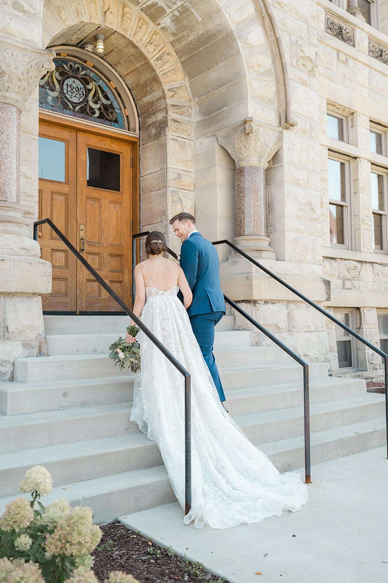 couple climbing concrete stairs to wooden doors with beautiful stained glass arch etched with 1893