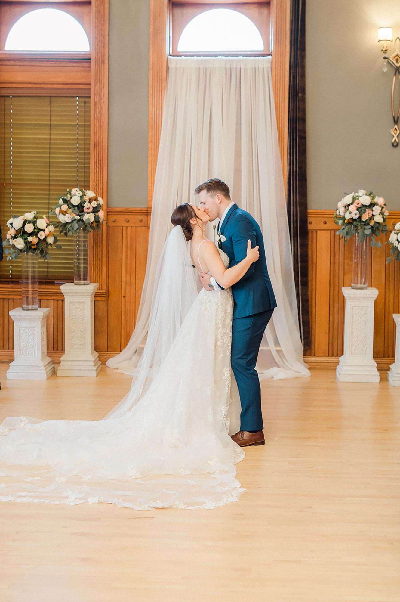 couple sharing a kiss at tulle altar, stone columns with clear hurricane vases and large centerpieces in background