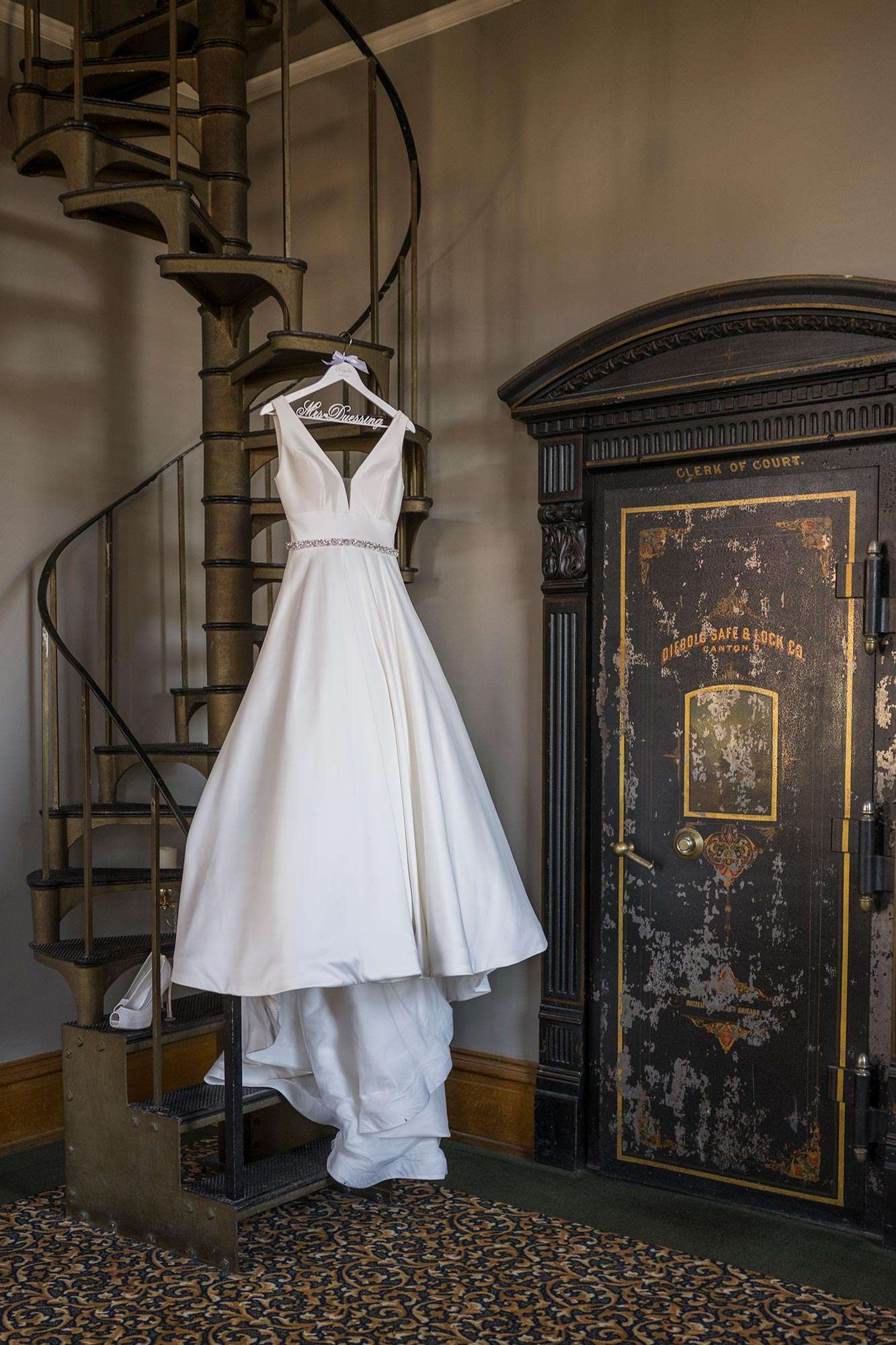 White satin bridal gown with rhinestone embellished waistline from Bliss hanging on monogrammed hanger on spiral staircase