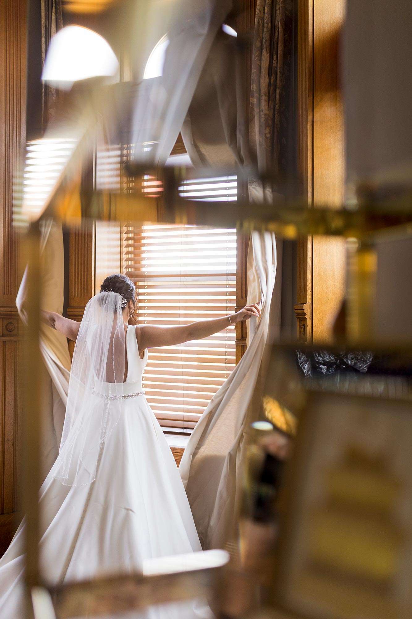 bride in gown and veil by Bliss looking out window while pulling back shades