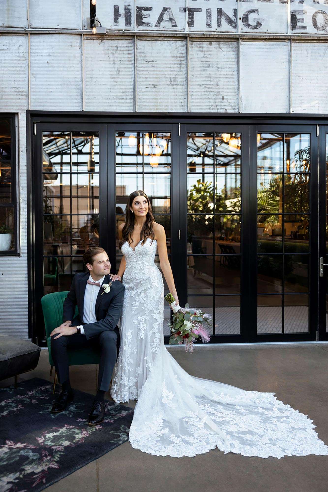 couple in front of black paned doors as groom sits on green chair couple in front of black paned doors as groom sits on green chair