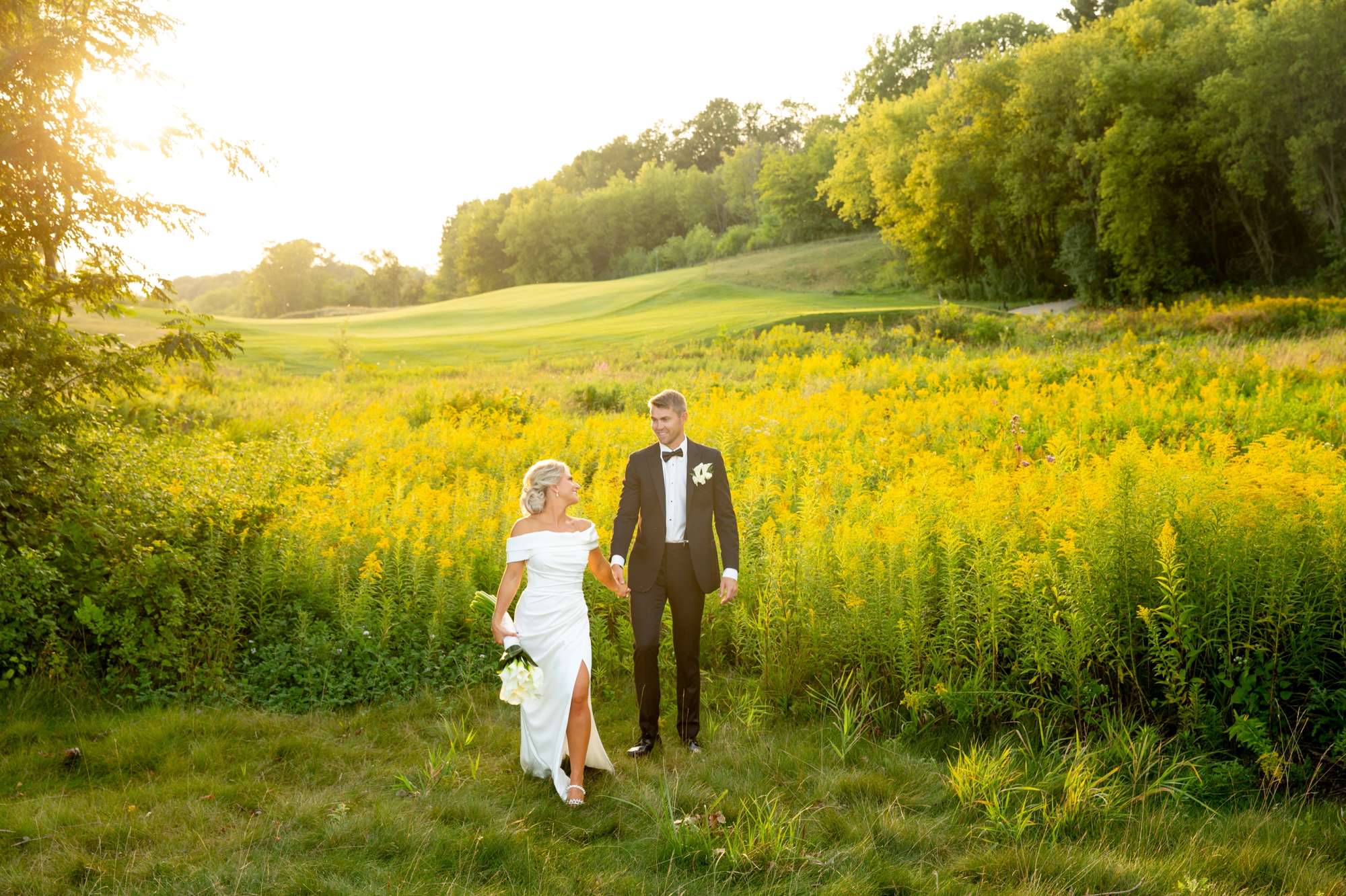 Bride in off the shoulder wedding dress and groom in black tuxedo as they walk through a field.