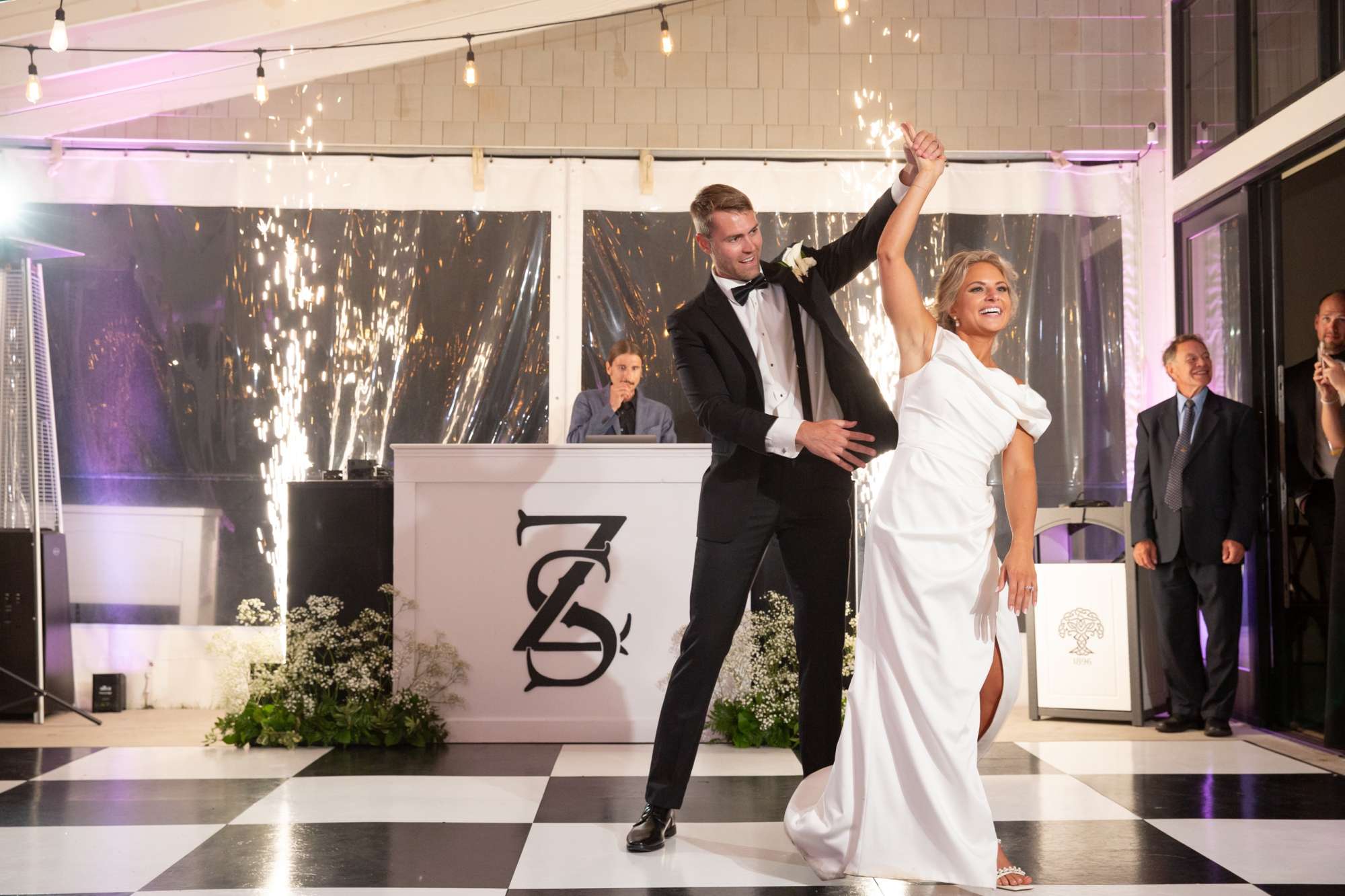 Groom twirling bride on checkered dance floor in front of custom logo and cold sparks.