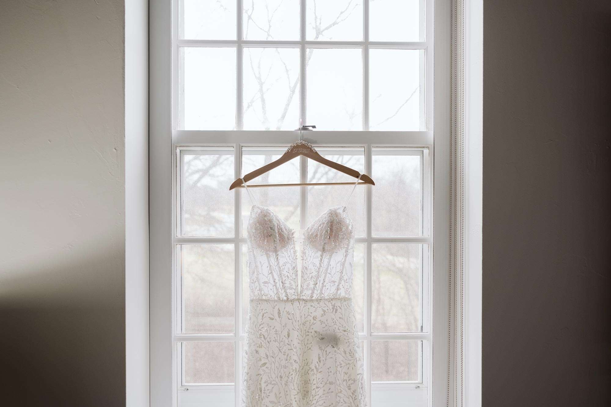 wedding gown with sheer boned bodice and shimmery vine like detail from Amelishan Bridal on hanger in front of white paned window Amelishan Bridal