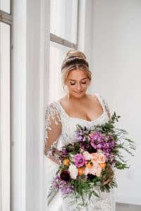 Bride with high bun and square neck dress looking down at large purple wedding bouquet. Bride with high bun and square neck dress looking down at large purple wedding bouquet.Bride with high bun and square neck dress looking down at large purple wedding bouquet.