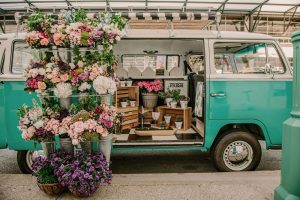 The side of a turquoise and white 1971 VW minibus is open, showcasing an enchanting display of colorful floral arrangements.