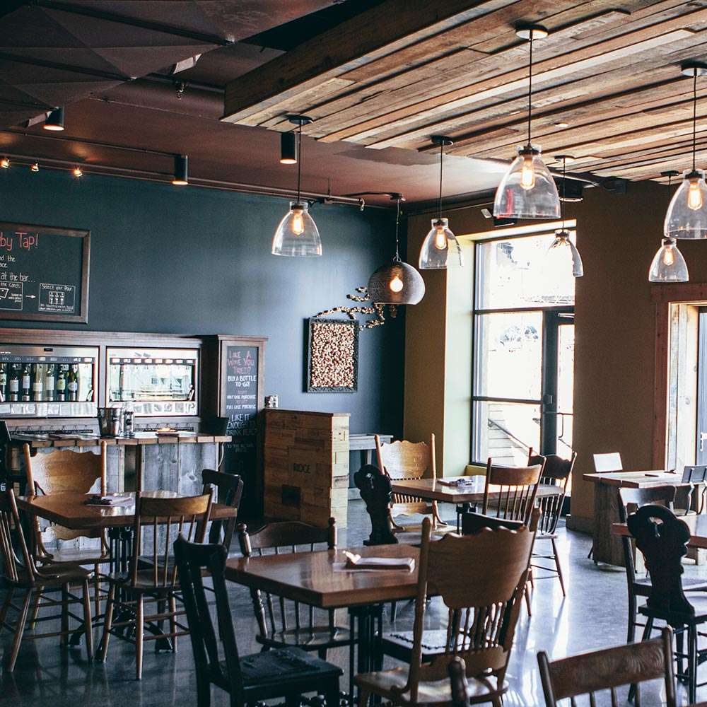 A cozy, rustic interior of The Ruby Tap wine bar featuring wooden tables and chairs, warm lighting from glass pendant lamps, and a wood-paneled ceiling.