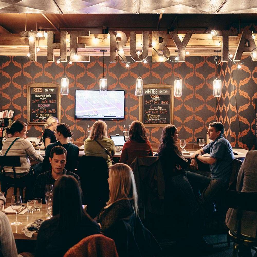A lively bar area at The Ruby Tap with people seated along a bar counter. The background has a patterned wallpaper, and the bar's name,