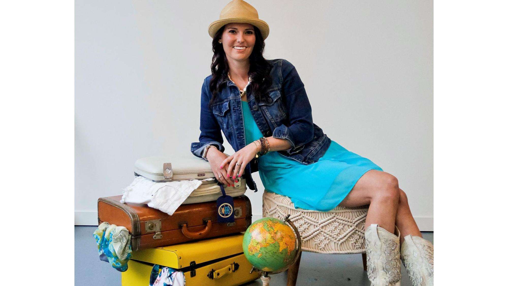 Travel_with_Becky_landscape Becky Jaskowiak in a blue dress, denim jacket, and cowboy boots sits next to a stack of vintage suitcases and a globe, showcasing the spirit of adventure and travel planning with Travel with Becky.