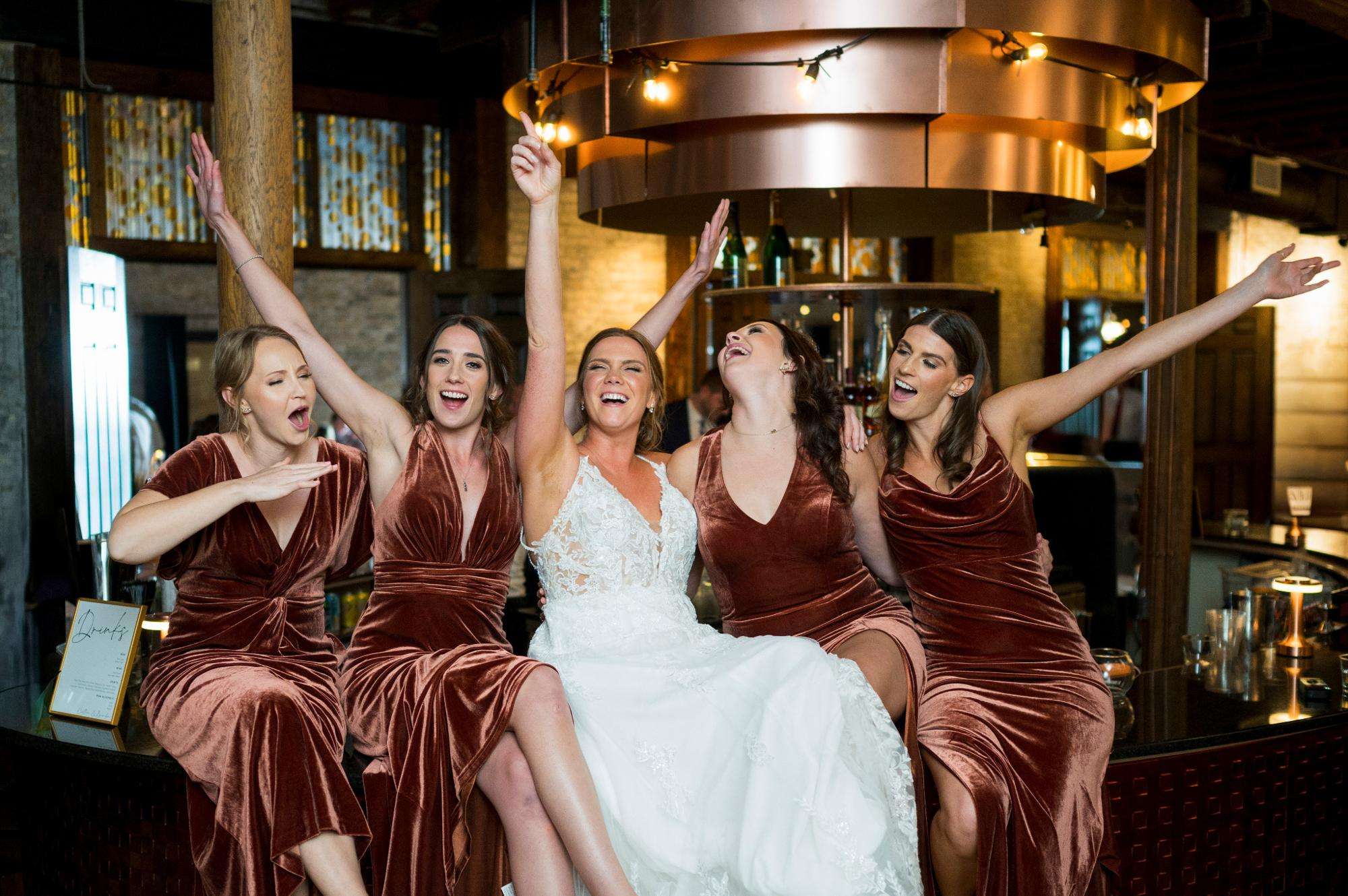 The bride and her bridesmaids burst with laughter and celebration while sitting on a stylish bar, captured by Allysha Noelle Photography.