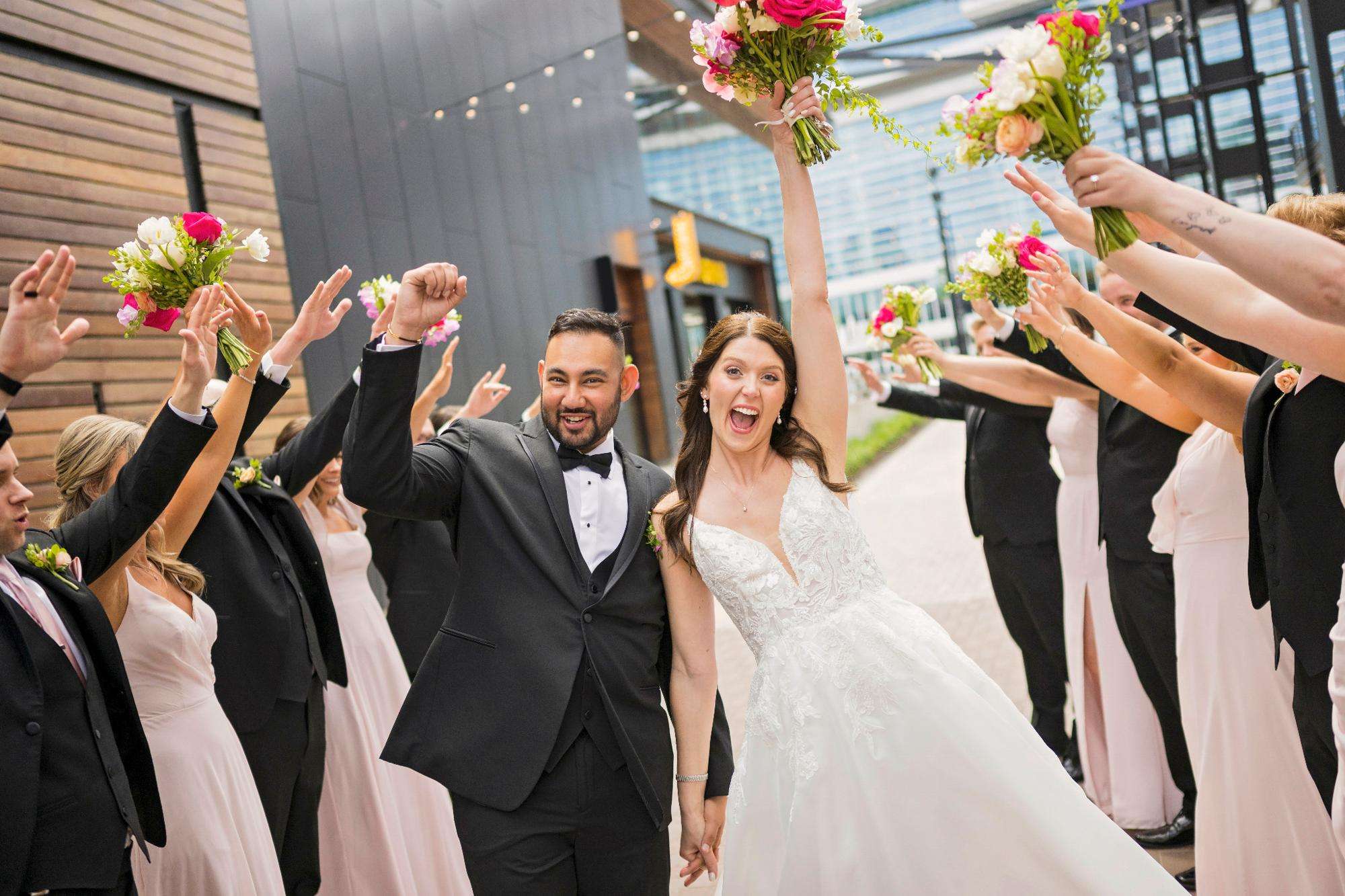 The newlyweds celebrate joyfully with their wedding party forming a floral tunnel, captured by Allysha Noelle Photography.