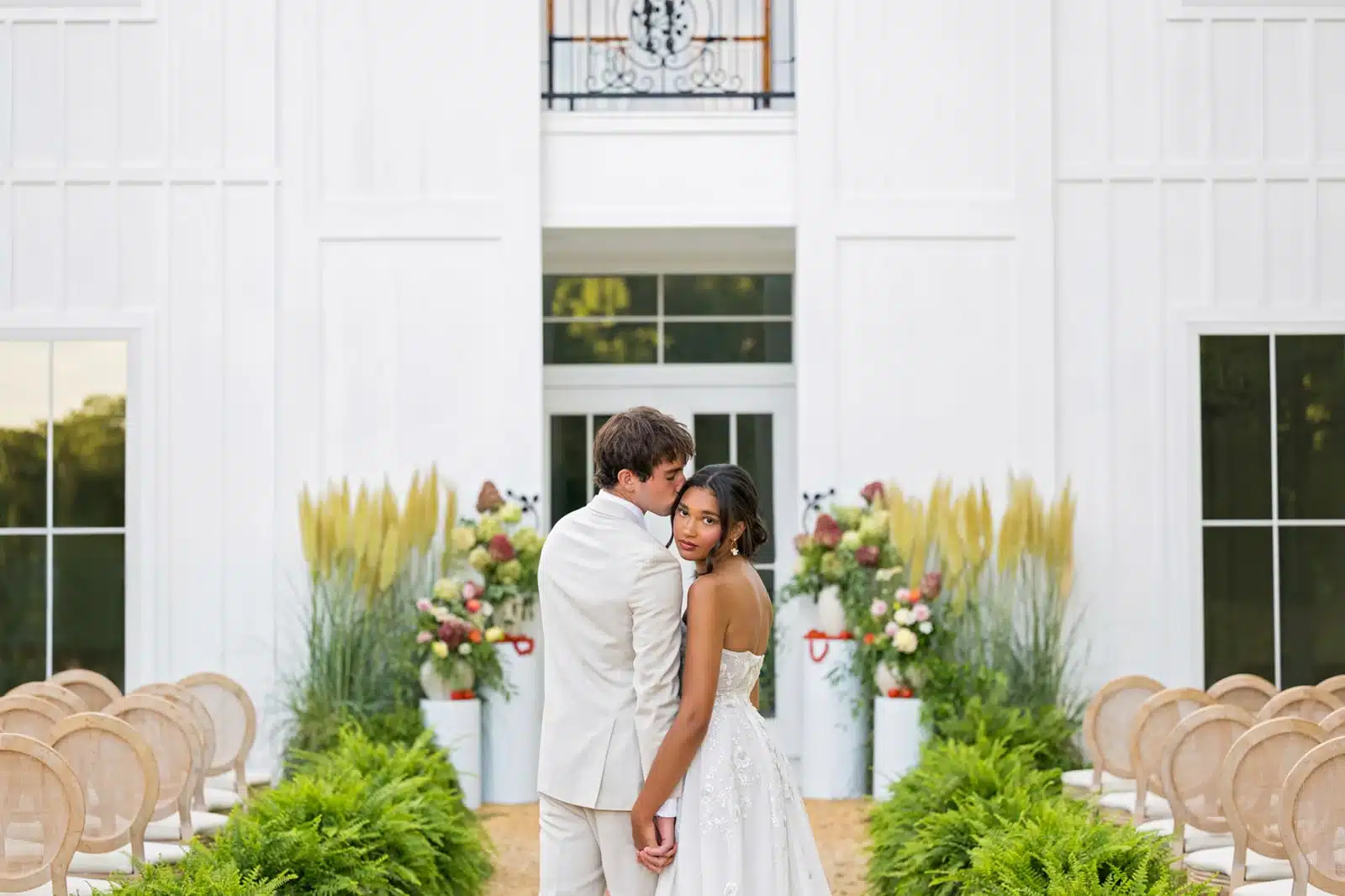 Bride gazing over her shoulder with flowers and white wedding venue in the background.