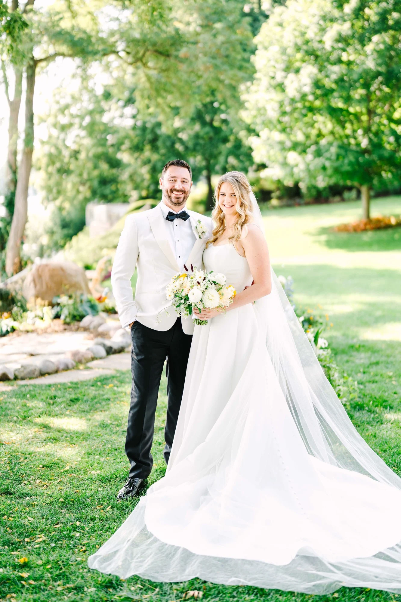 Bride and groom post on their lawn in Lake Geneva with her dress sweeping to the side and her cathedral veil. Bride and groom post on their lawn in Lake Geneva with her dress sweeping to the side and her cathedral veil.