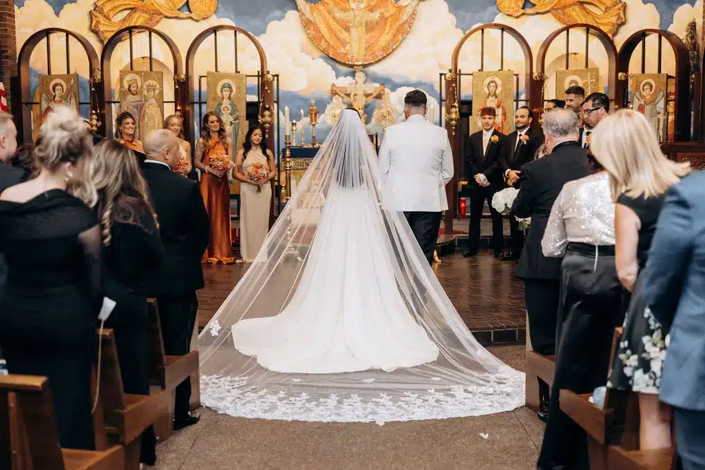 View of bride and groom at their wedding ceremony at Sts Constantine & helen with brides long cathedral veil is draped along the aisle. View of bride and groom at their wedding ceremony at Sts Constantine & helen with brides long cathedral veil is draped along the aisle.