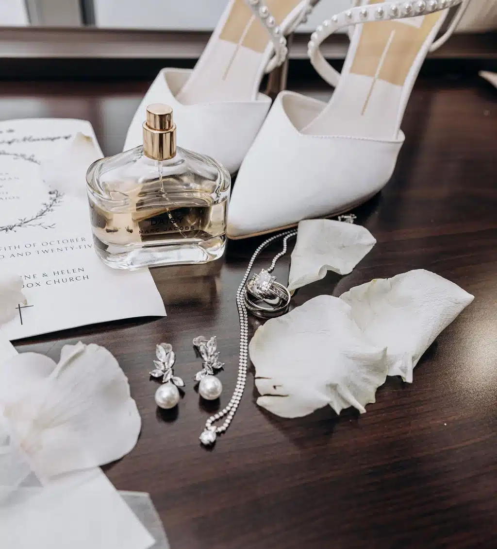 Brides wedding details of white, pointed shoes, perfume, drop chandelier earrings, invitation and diamond necklace. Brides wedding details of white, pointed shoes, perfume, drop chandelier earrings, invitation and diamond necklace.