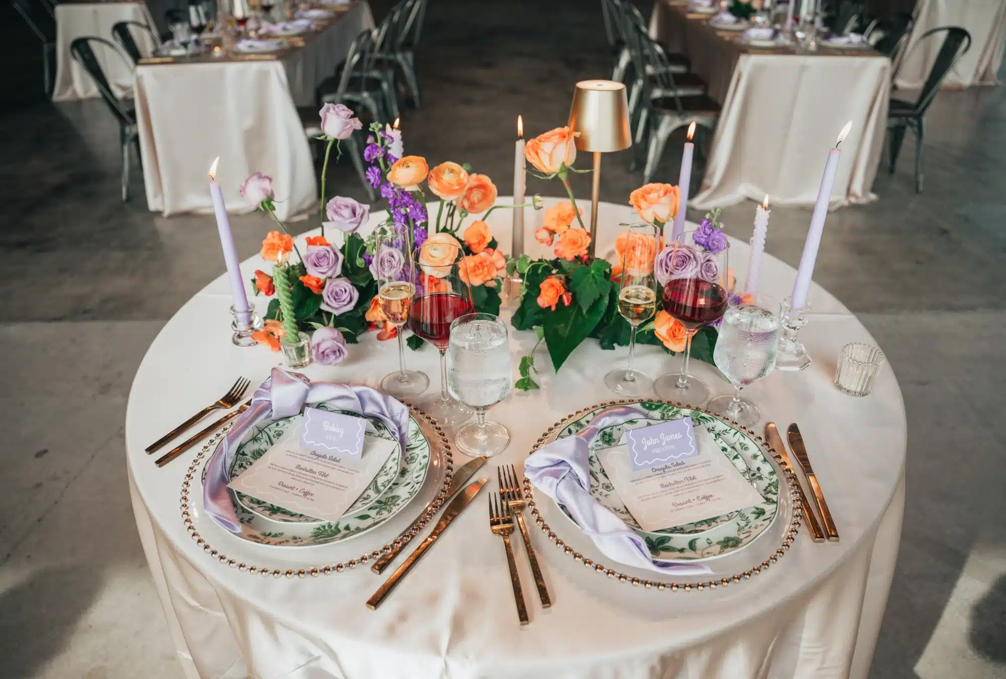 Sweetheart table at Jan Serr Studio with gold tabletop lamp, florals of purple and orange, gold beaded chargers, green floral plates and gold flatware. Sweetheart table at Jan Serr Studio with gold tabletop lamp, florals of purple and orange, gold beaded chargers, green floral plates and gold flatware.