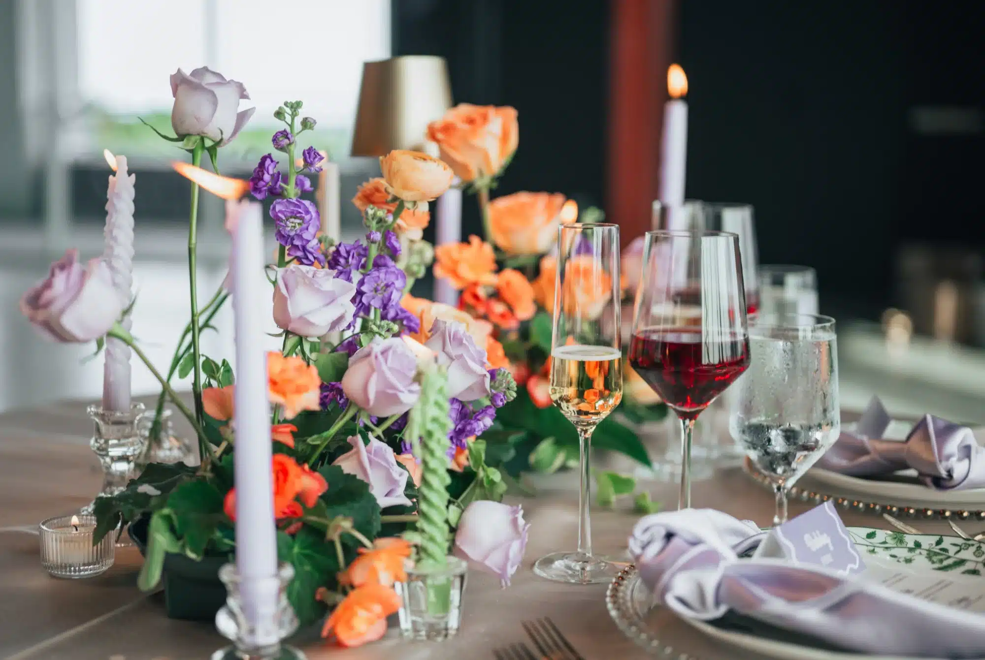 Details of tabletop setup in the colors of purple, orange and coral with purple taper candles and wine glasses with gold beaded chargers. Details of tabletop setup in the colors of purple, orange and coral with purple taper candles and wine glasses with gold beaded chargers.