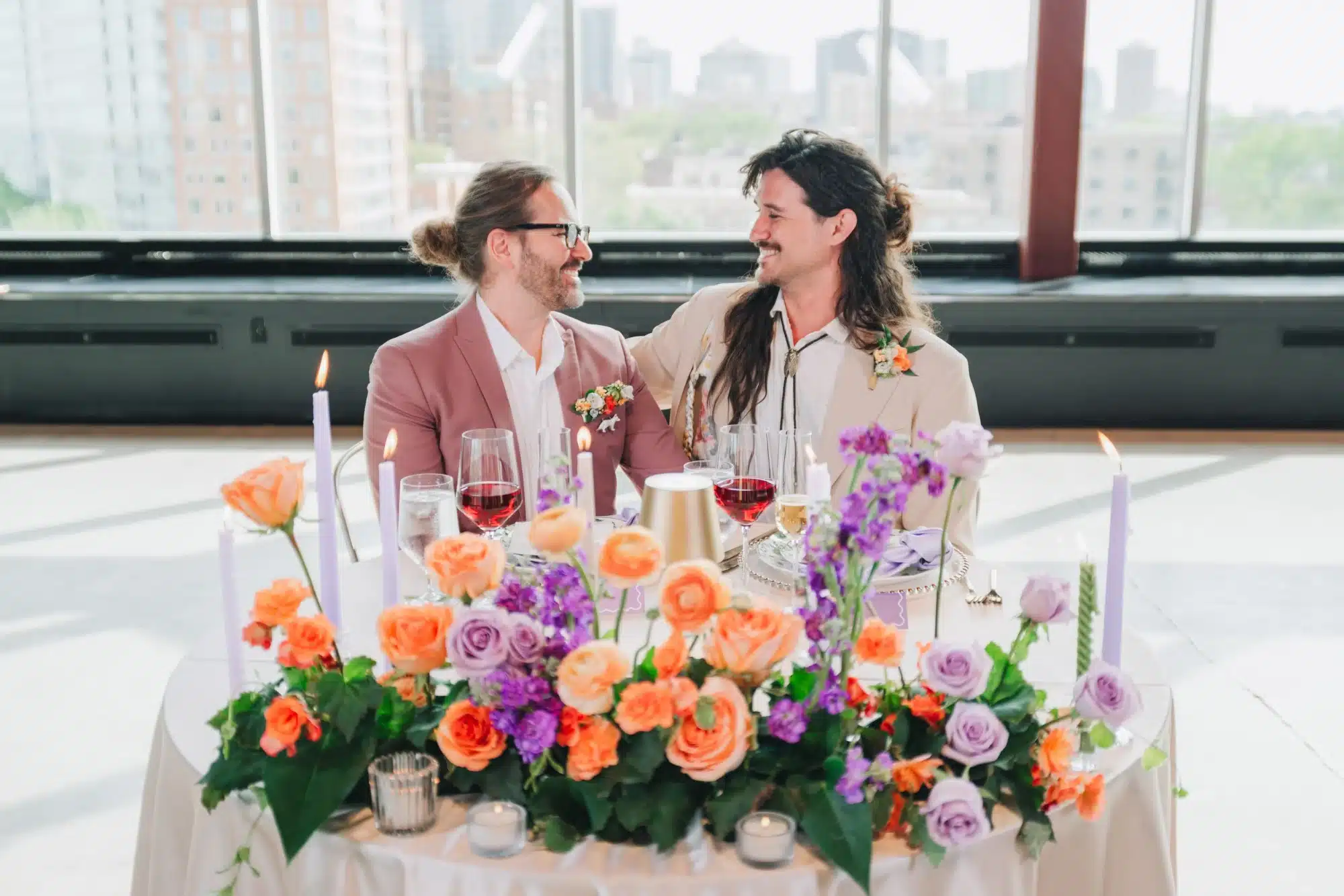 Grooms sit at sweetheart table at Jan Serr Studio, table filled with purple, orange and coral flowers and purple taper candles. Grooms sit at sweetheart table at Jan Serr Studio, table filled with purple, orange and coral flowers and purple taper candles.