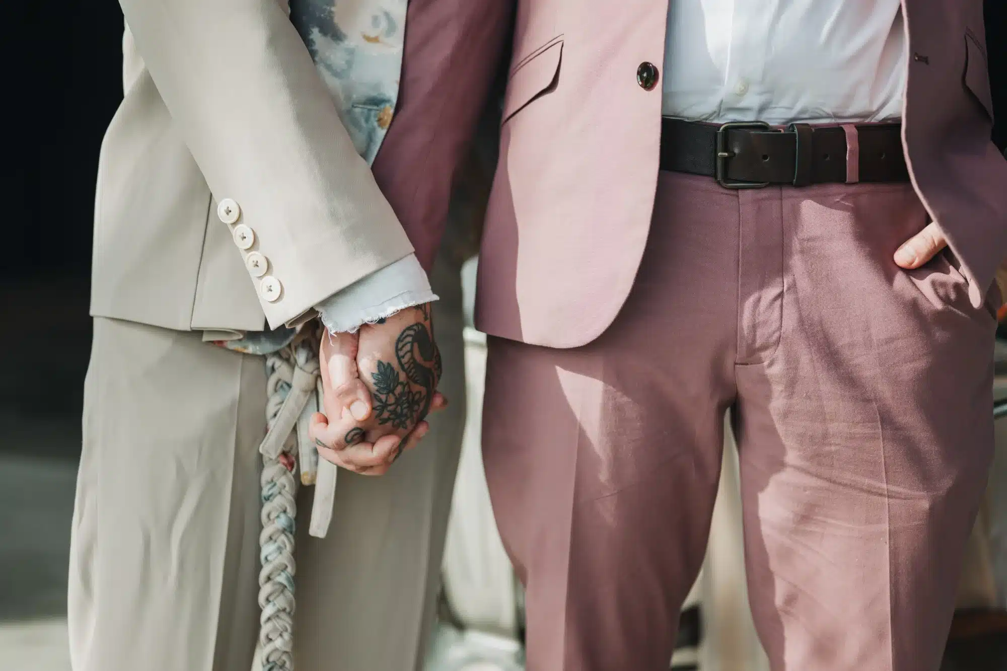 Close up photo of grooms holding hands in tan and mauve suits. Close up photo of grooms holding hands in tan and mauve suits.
