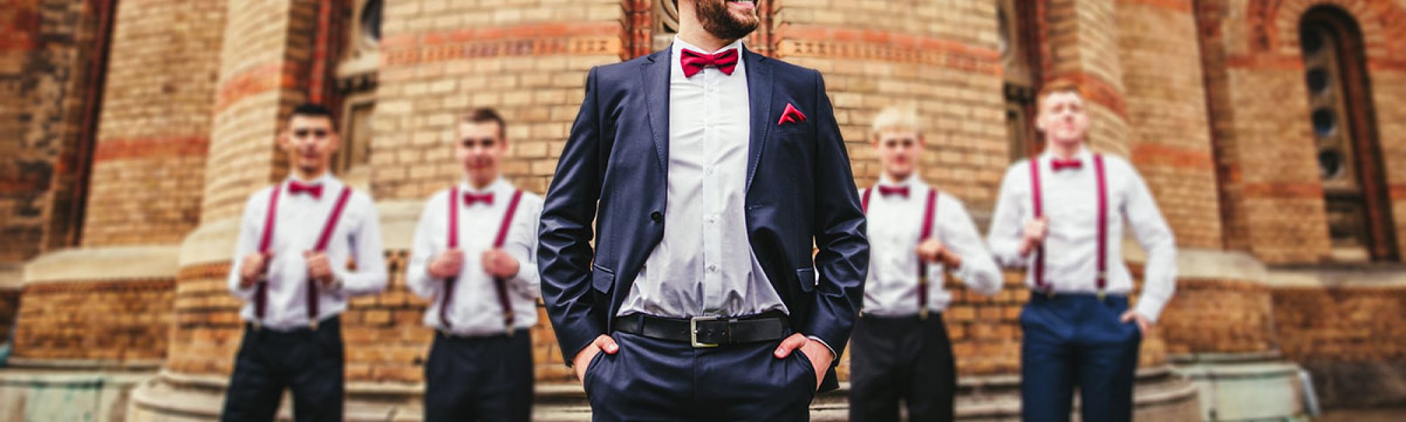 groom with red bowtie standing in front of groomsmen wearing suspenders