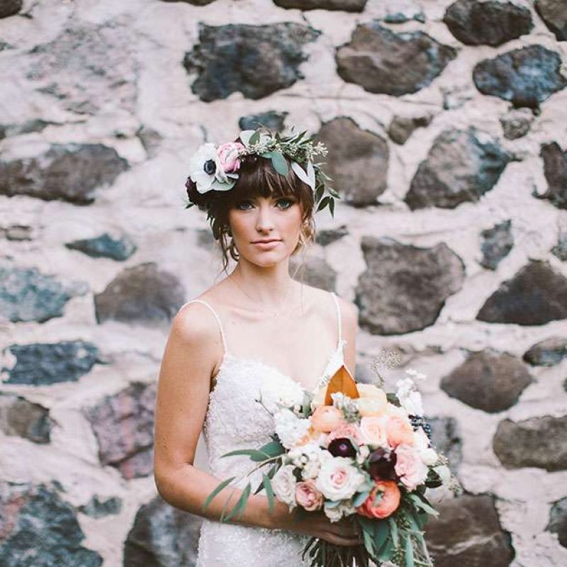 Bride with bouquet and floral wreath poses in front of stone wall