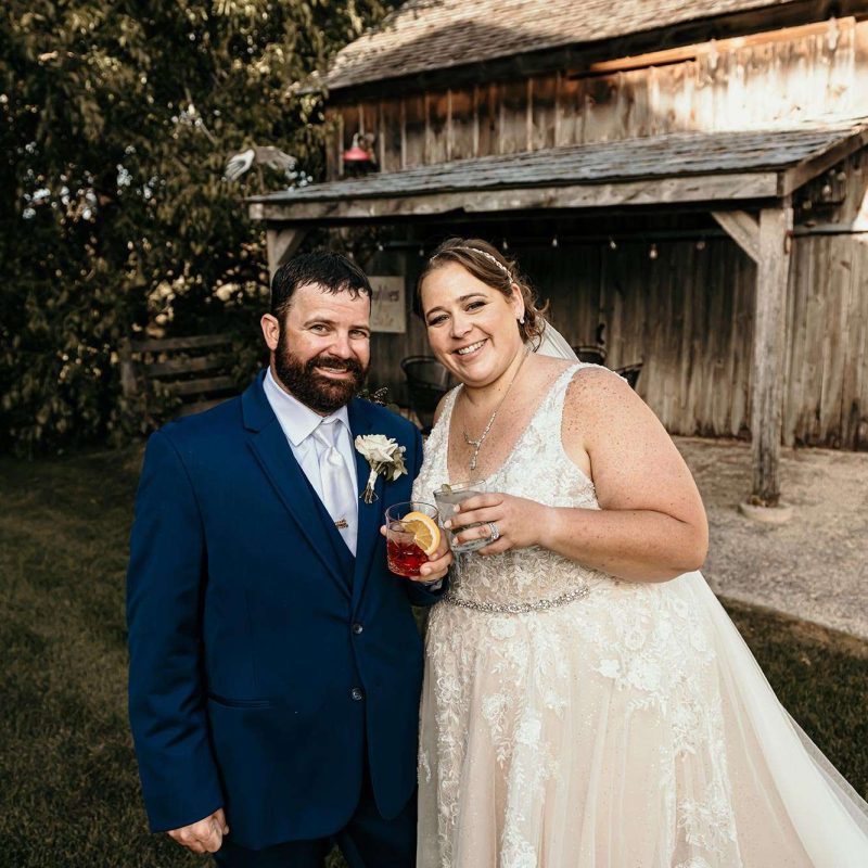 couple outside rustic barn toasting with signature drinks in hand