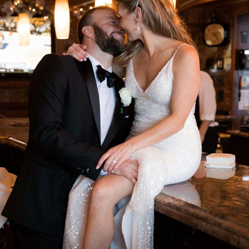 Bride sitting on top of bar while couple shares a kiss
